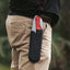 Person wearing a black leather sheath with a red pocket knife on beige pants against a blurred natural background