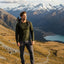 A man stands on a rocky mountain ridge wearing an olive green long-sleeve shirt, dark gray hiking pants, and brown hiking boots, with a backpack and a scenic mountain landscape with a lake in the background.