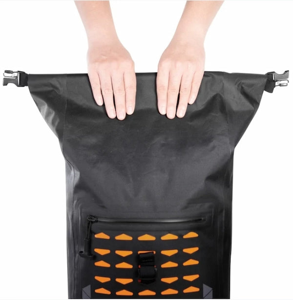 A black waterproof roll-top bag with a front zip pocket and orange vent-like accents, held open by two hands against a white background.