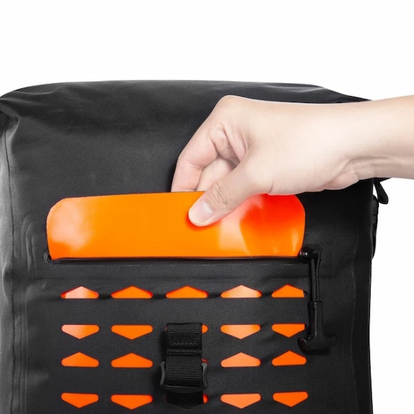 A close-up of a black backpack with an orange waterproof flap pocket being opened by a hand, showing the interior drainage holes below.