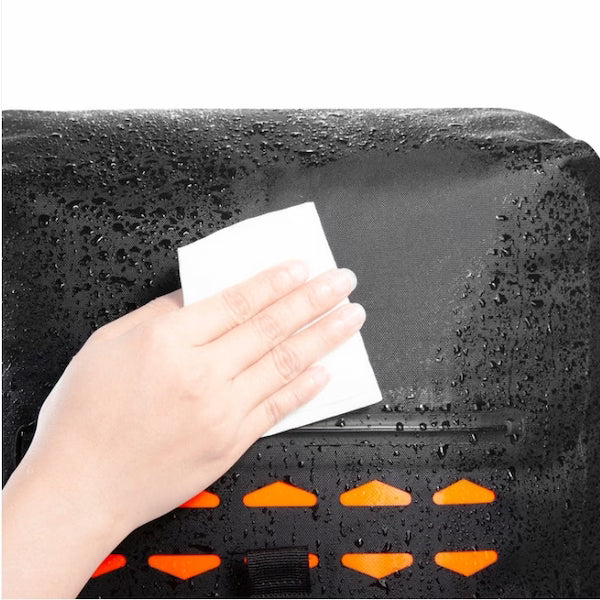 A close-up of a hand holding a white card against a black waterproof fabric surface covered in water droplets, with orange accents visible along the bottom edge.