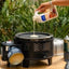 Person pouring milk into a portable stove with a cup, surrounded by outdoor elements.