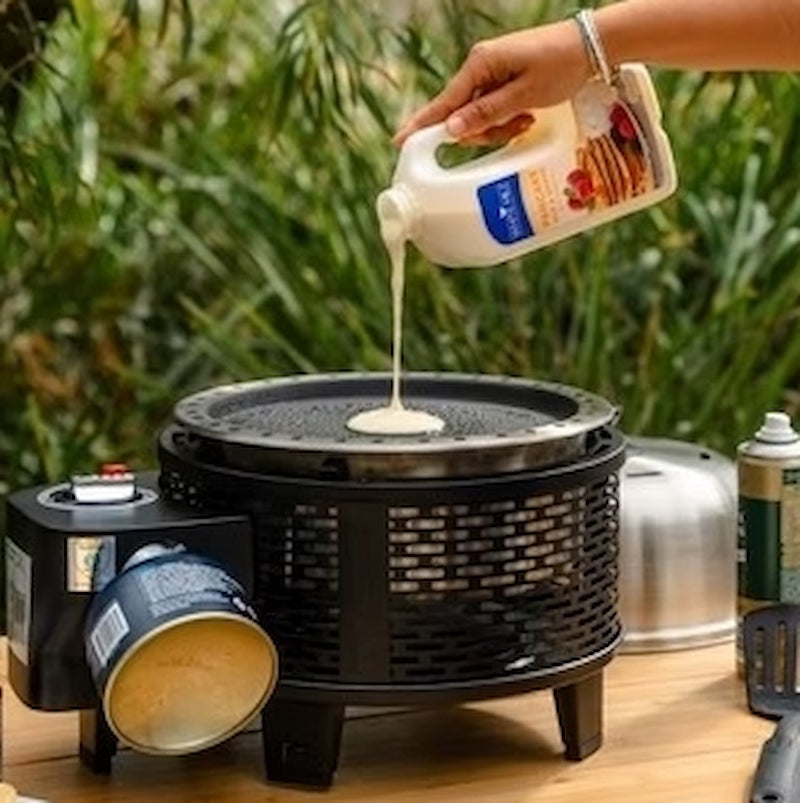 Person pouring milk into a portable stove with a cup, surrounded by outdoor elements.