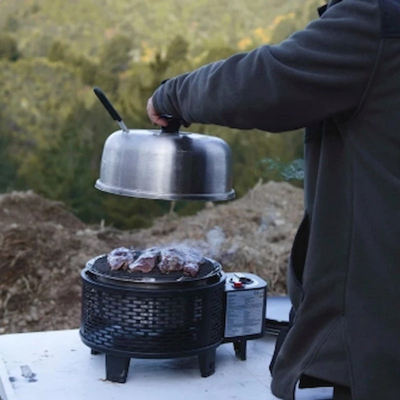 Person cooking on a portable grill with a metal lid outdoors