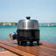 Portable grill on a wooden surface with a scenic background of water and sky.