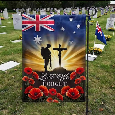 A memorial garden flag displayed on a black metal stand in a grassy cemetery. The flag features the Australian flag at the top, a silhouette of a soldier facing a cross with a radiant yellow background, red poppy flowers at the bottom, and the text 'Lest We Forget'.