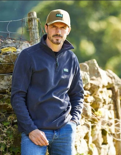 Man wearing a navy fleece pullover with a logo, standing outdoors near a stone wall.