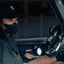 Person inside a vehicle holding a flashlight, wearing a black cap and mask.