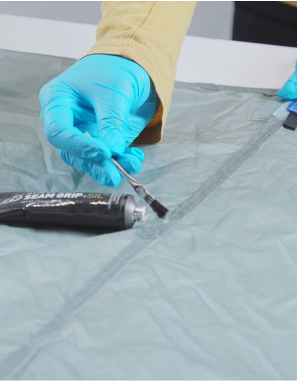 Person applying Seam Grip SIL sealant to a zipper with blue gloves on a gray surface.