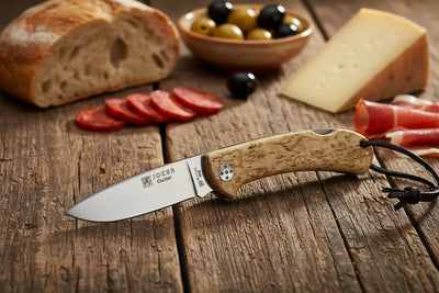 Knife with wooden handle on a wooden surface with bread, olives, and cheese in the background