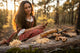 Woman in traditional attire sitting in a forest with a knife and wooden tools on a log.