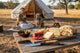 Woman with a dog by a tent in a field, with a wooden table displaying food and a knife.
