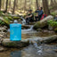 Blue water bottle on a rock in a forest setting with people in the background.