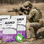 A soldier in camouflage kneels outdoors holding a rifle, with three bags of ALOPEX Airsoft Precision pellets in the foreground. The bags are white and purple with the ALOPEX brand name and '0.