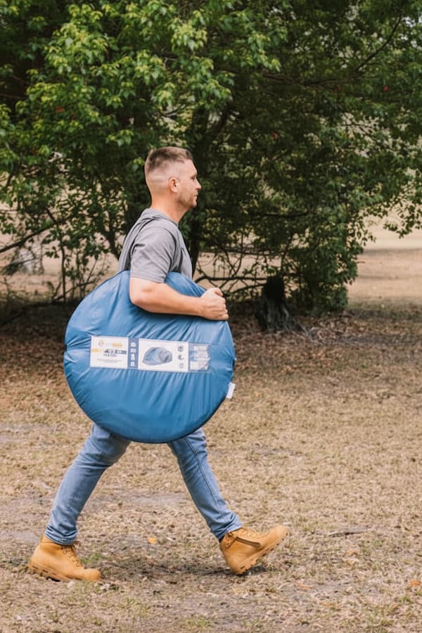 A man in a gray t-shirt, blue jeans, and tan boots walks outdoors carrying a large blue circular camping tent bag with printed white labels.