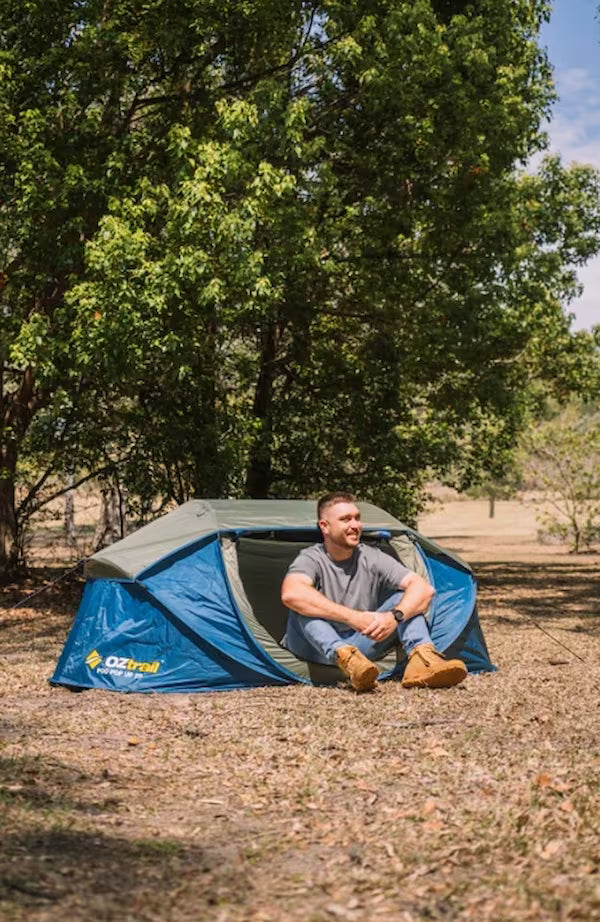 Man sitting next to a blue tent in a natural setting with trees and open space.