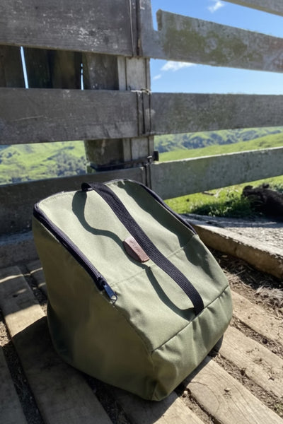 An olive green insulated cooler bag with black zipper and strap details, resting on a weathered wooden fence outdoors with a scenic green hillside in the background.