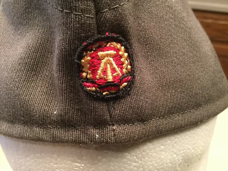 Close-up of a gray cap with a embroidered emblem featuring red, gold, and black colors.